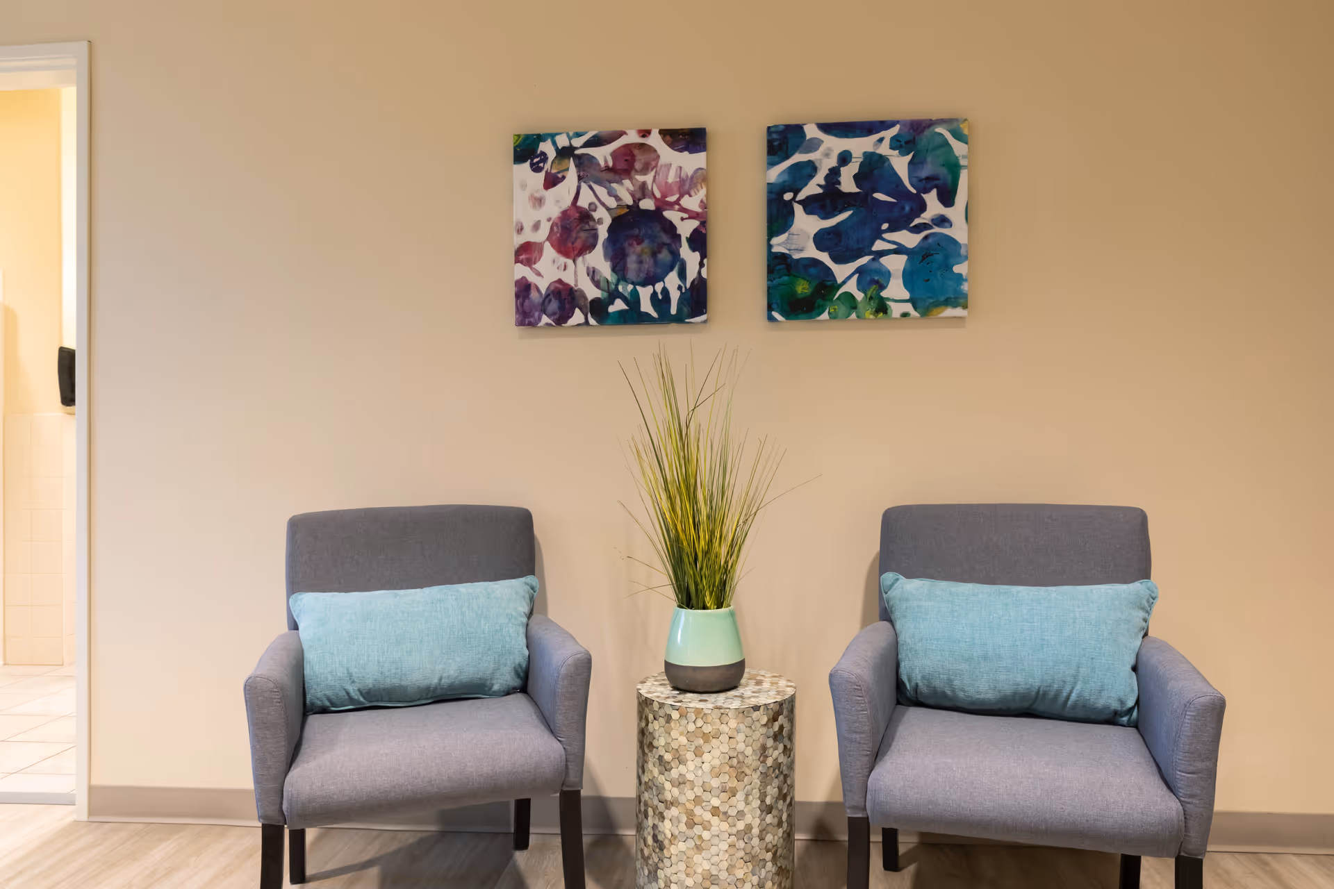 Two gray armchairs with light blue cushions are placed on either side of a small round table with a decorative plant in a vase. Above the chairs, there are two colorful abstract paintings on a beige wall. A doorway to a tiled room is visible on the left side.