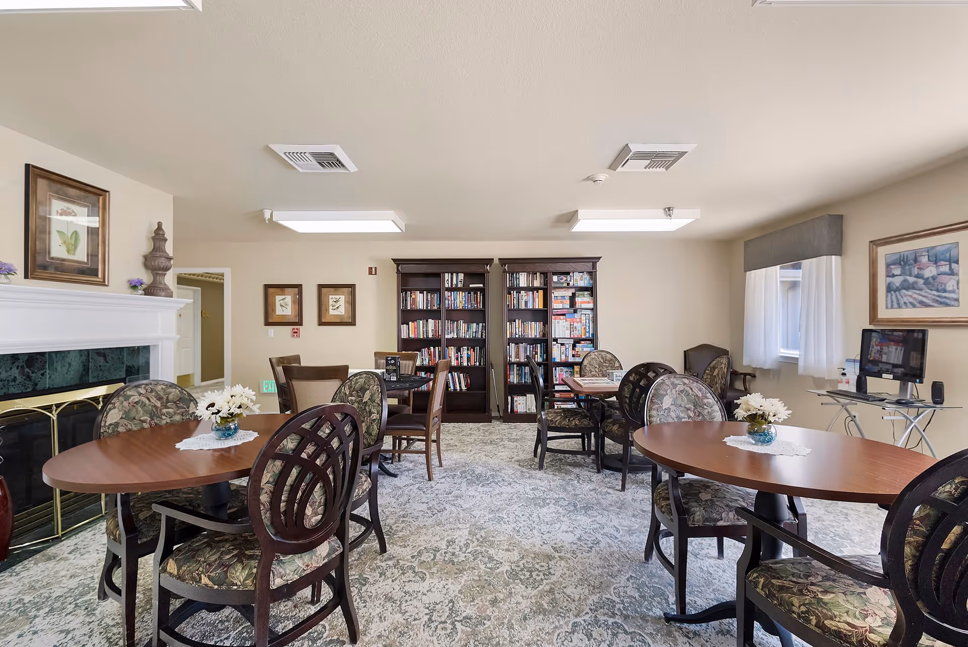 A cozy common room with multiple round wooden tables and floral upholstered chairs. There are two large bookshelves filled with books and board games against the far wall. The room has a fireplace on the left side with decorative items on the mantle, framed artwork on the walls, and a window with white curtains on the right. A computer workstation is set up near the window.