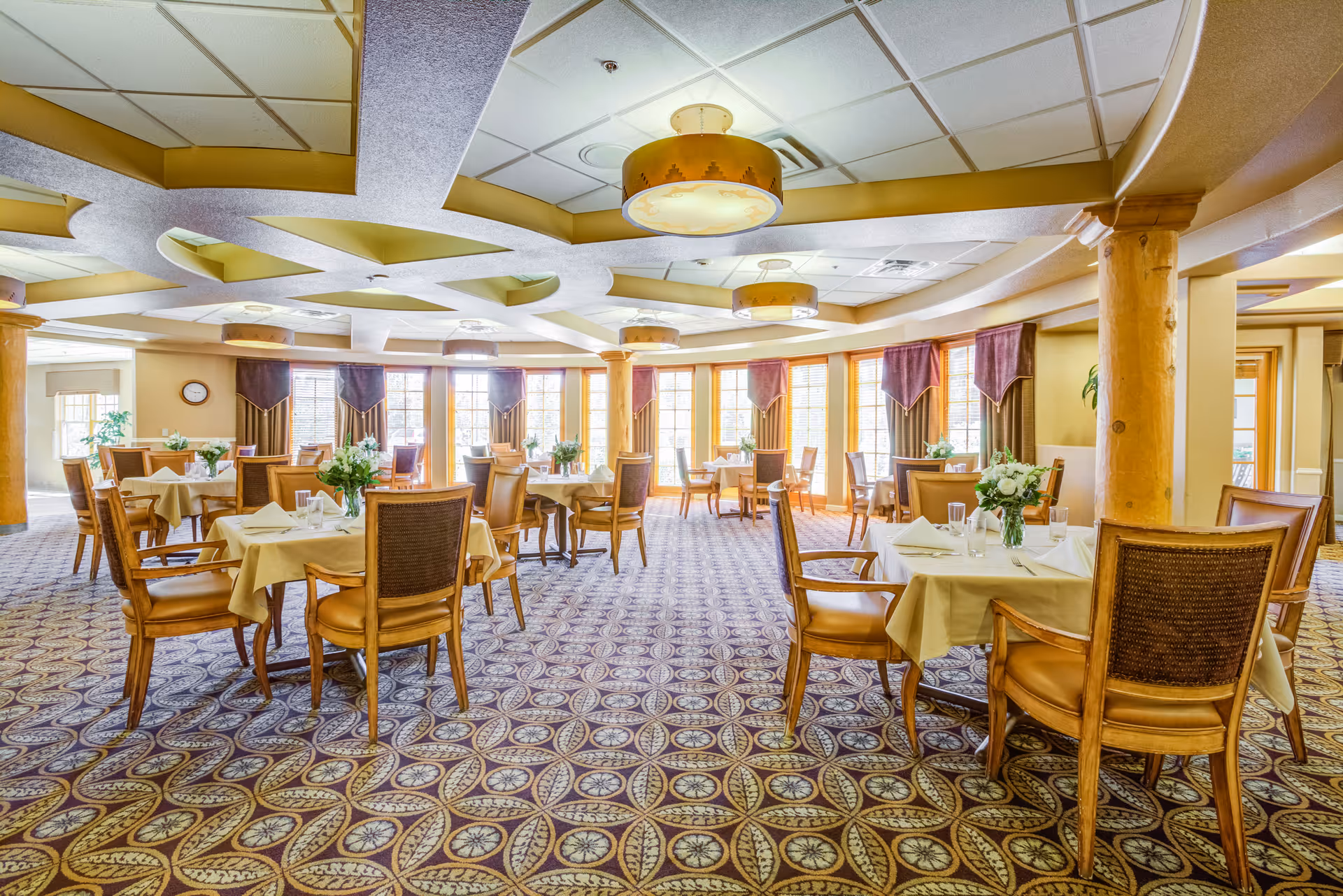 A spacious dining room in a senior living community with multiple round tables covered with beige tablecloths, each set with glasses, napkins, and floral centerpieces. The room features large windows with brown and purple curtains, wooden chairs with cushioned seats, patterned carpet, and decorative ceiling lights.