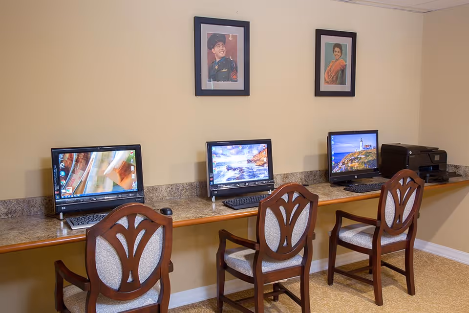 A row of three desktop computers and wooden chairs along a countertop in a communal computer room with framed portraits on the wall.