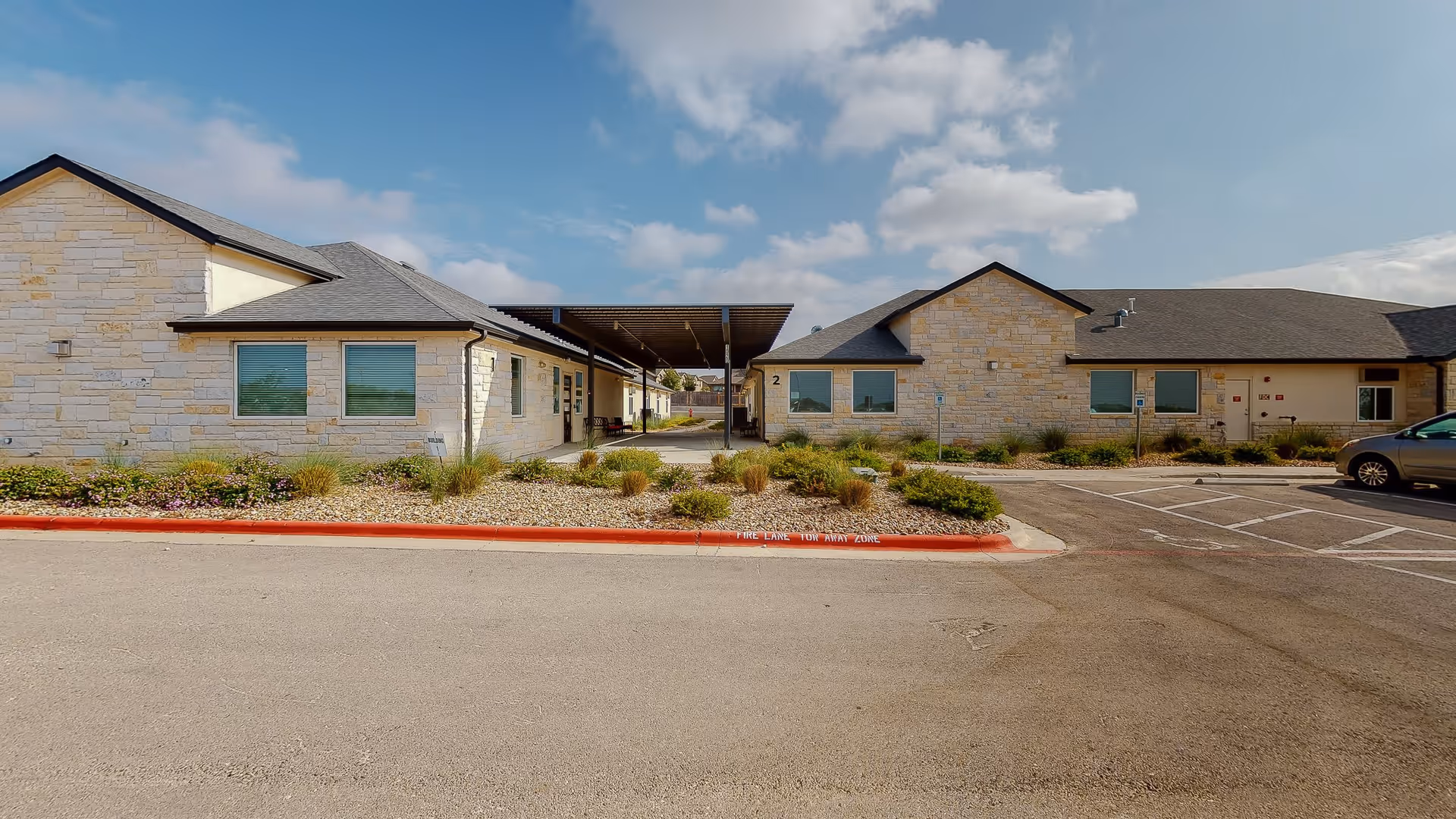 Exterior view of a single-story assisted living facility building with light-colored stone walls, multiple windows with blinds, a covered walkway between two sections of the building, landscaped bushes and rocks in front, and a parking area with a car on the right side under a partly cloudy sky.