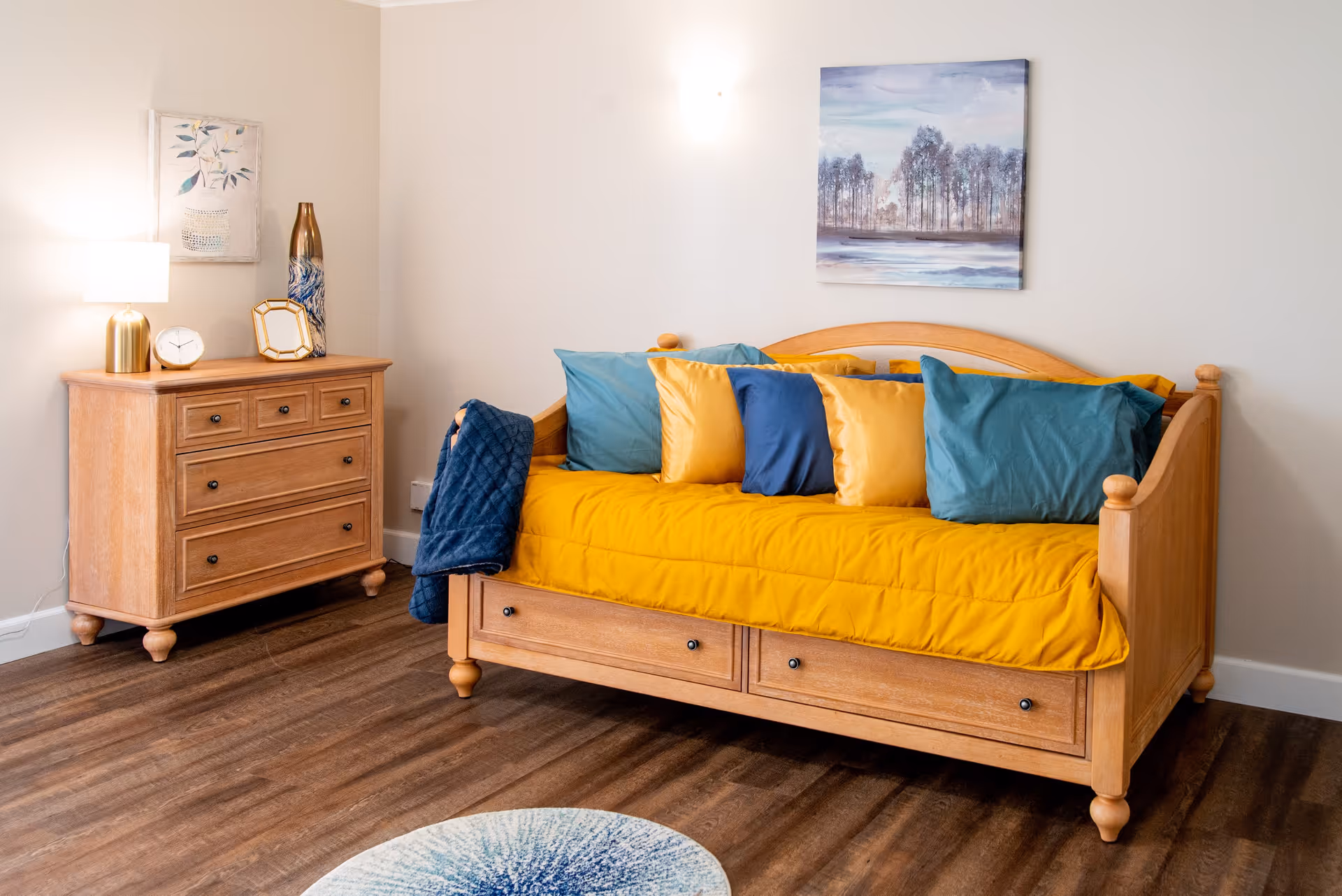 A cozy room with a wooden daybed covered with a bright yellow quilt and multiple blue and yellow pillows. Next to the daybed is a wooden dresser with a lamp, clock, decorative vase, and a framed picture. The walls are light-colored with two pieces of artwork hanging, and the floor is wooden with a round blue and white rug partially visible.