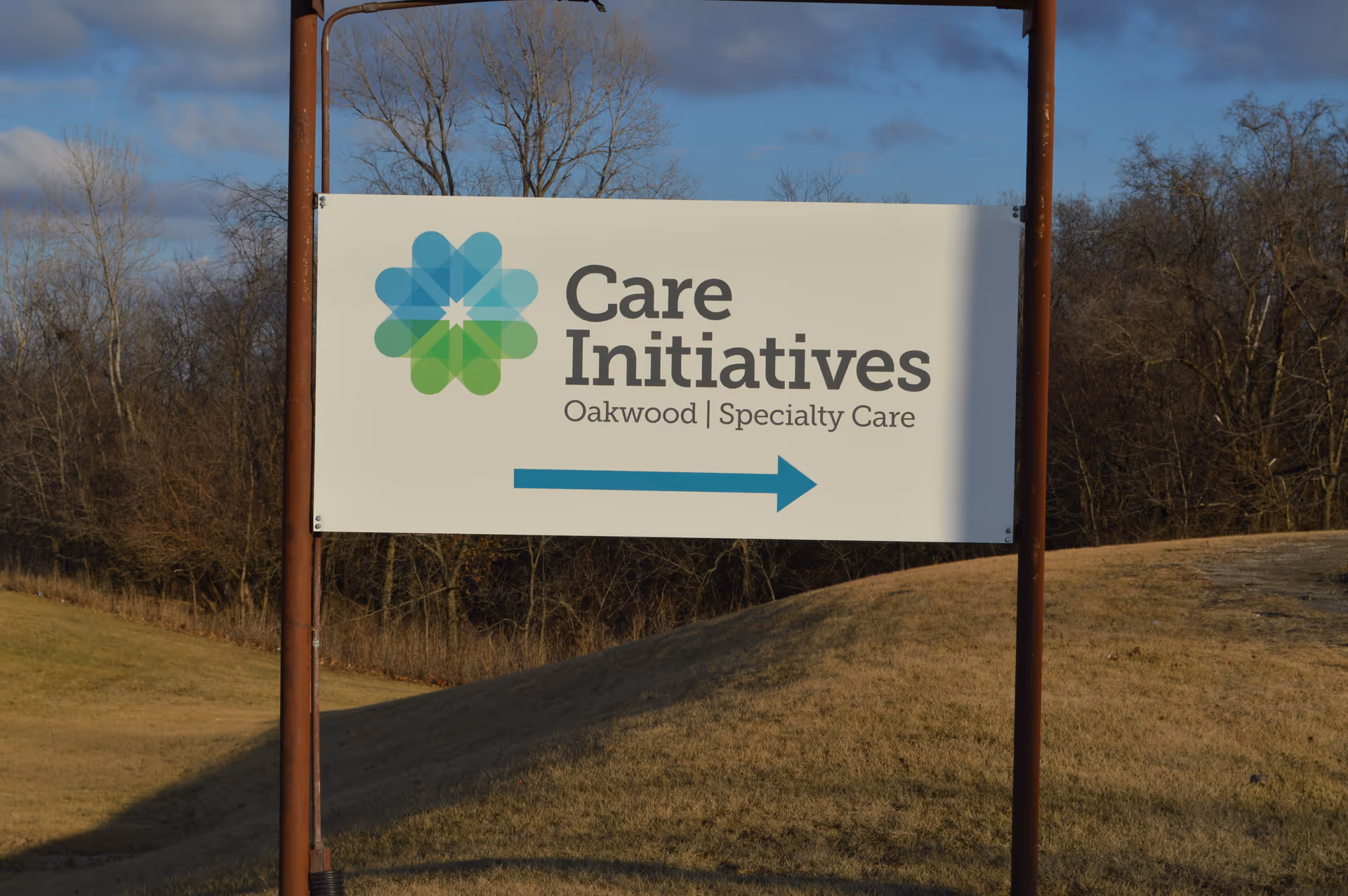 Outdoor sign for Care Initiatives Oakwood Specialty Care with a blue and green logo and a blue arrow pointing to the right, set against a background of grass and leafless trees under a partly cloudy sky.