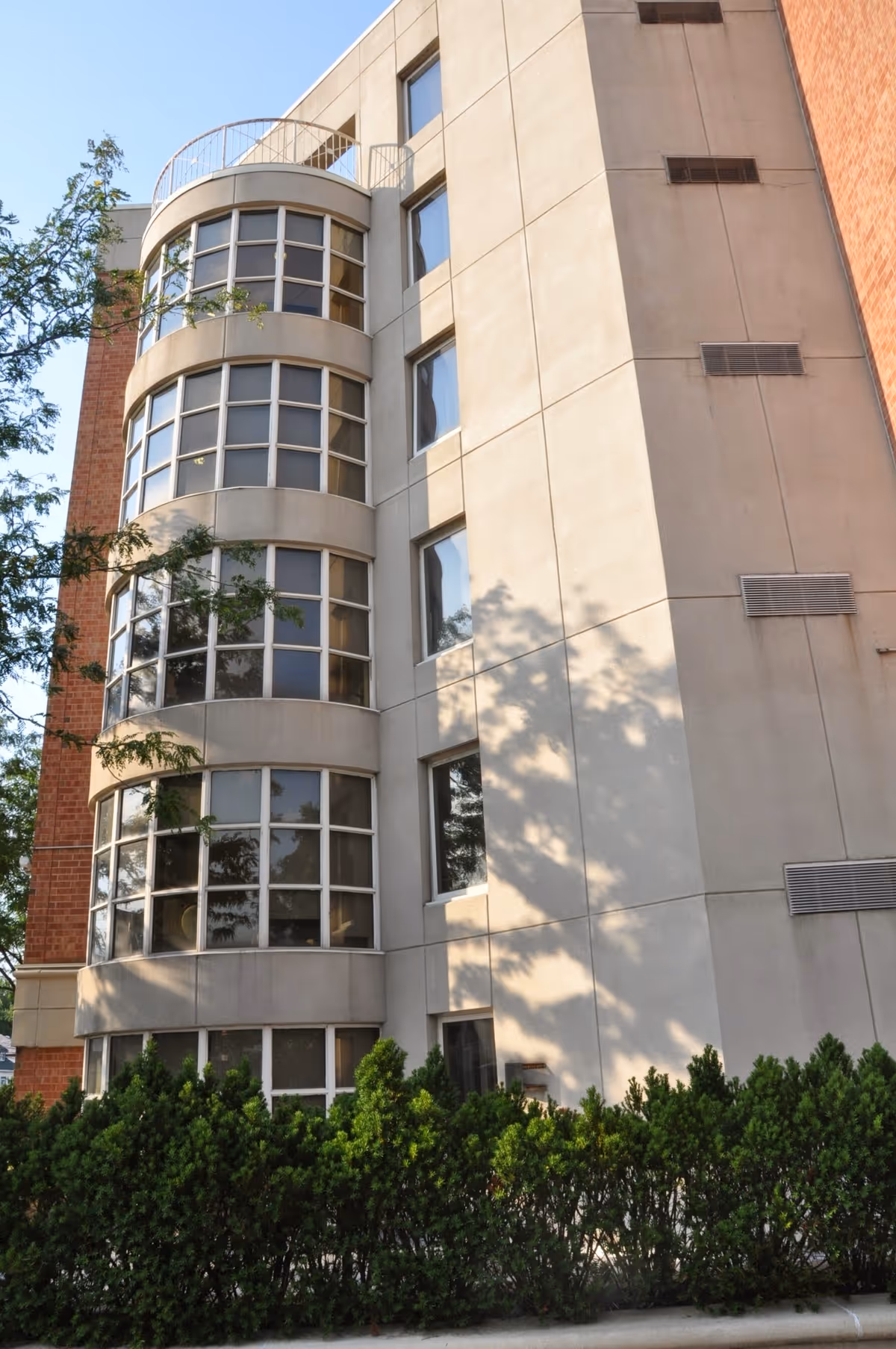 Exterior view of a multi-story building with large curved windows and beige walls, surrounded by green bushes and trees under a clear blue sky.