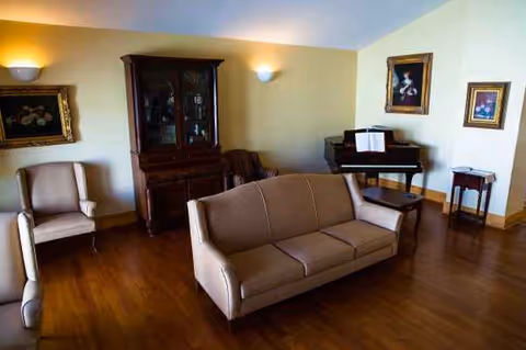 Cozy senior living living room with beige sofa, chairs, piano, and framed artwork.