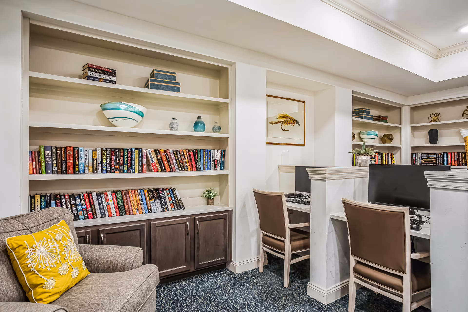 Cozy common area with built-in bookshelves, a cushioned chair with a yellow pillow, and two small computer workstations.
