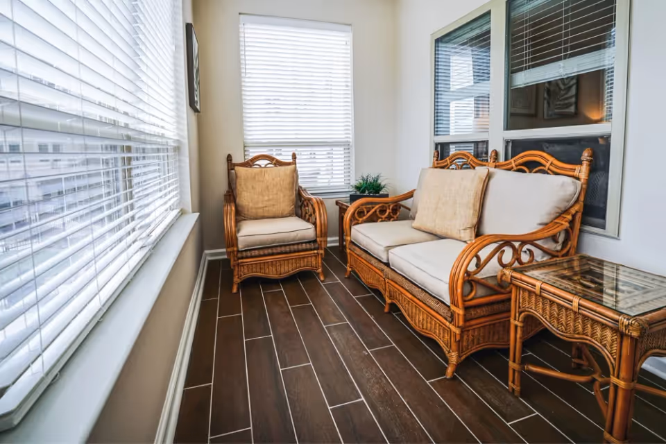 A bright indoor seating area with wicker furniture including a cushioned armchair, a cushioned loveseat, and a glass-top side table. The room has large windows with white blinds and dark wood-look tile flooring.
