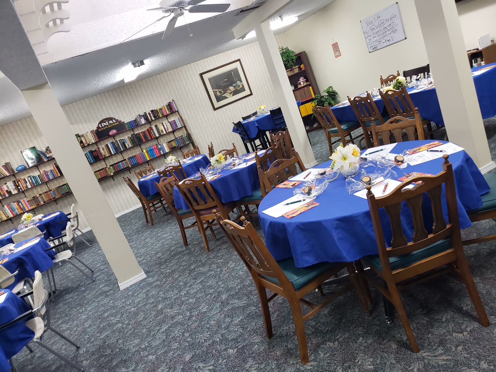 A room with multiple round tables covered in blue tablecloths, each set with papers, pens, and small flower arrangements. Wooden chairs and some white chairs surround the tables. Bookshelves filled with books line the walls, and a whiteboard with writing is visible on one wall. The room has carpeted flooring and ceiling fans.