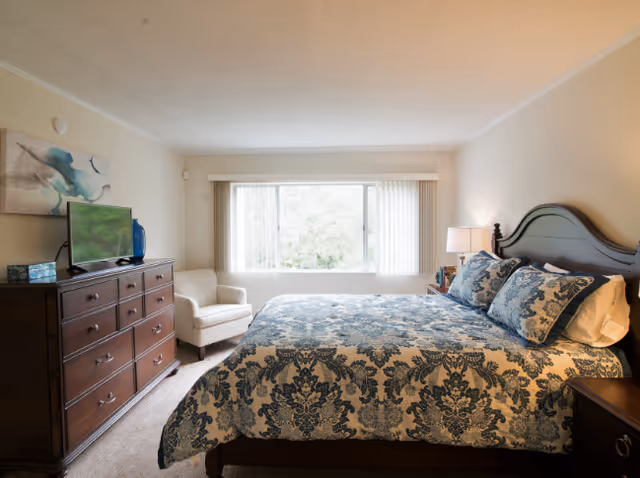 A well-lit bedroom featuring a large bed with a blue and white patterned comforter and matching pillows. To the right of the bed is a nightstand with a lamp and some decorative items. On the left side of the room, there is a wooden dresser with a flat-screen TV on top and a blue vase beside it. A comfortable armchair is positioned near a large window with vertical blinds, allowing natural light to fill the room.