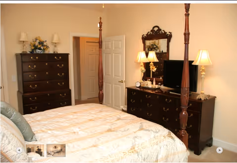 A bedroom with a large bed covered in a cream-colored quilt. The room features dark wooden furniture including a tall chest of drawers and a dresser with a mirror. Two table lamps with cream-colored shades are on the dresser, along with a small clock and decorative items. The walls are light-colored, and there is an open door leading to another room.