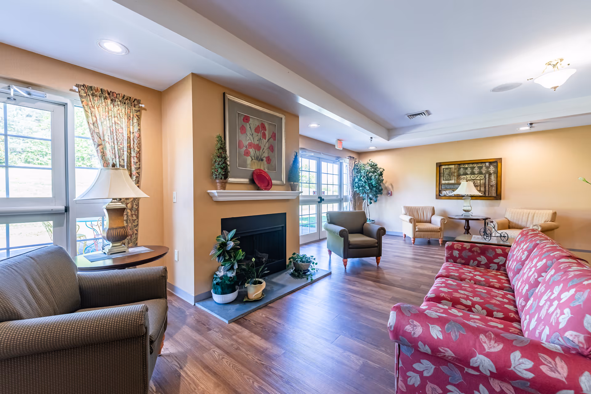 A bright and spacious living room in an assisted living facility featuring a red floral-patterned sofa, several armchairs, a fireplace with plants and a framed floral painting above it, wooden flooring, and large windows with floral curtains allowing natural light to fill the room.