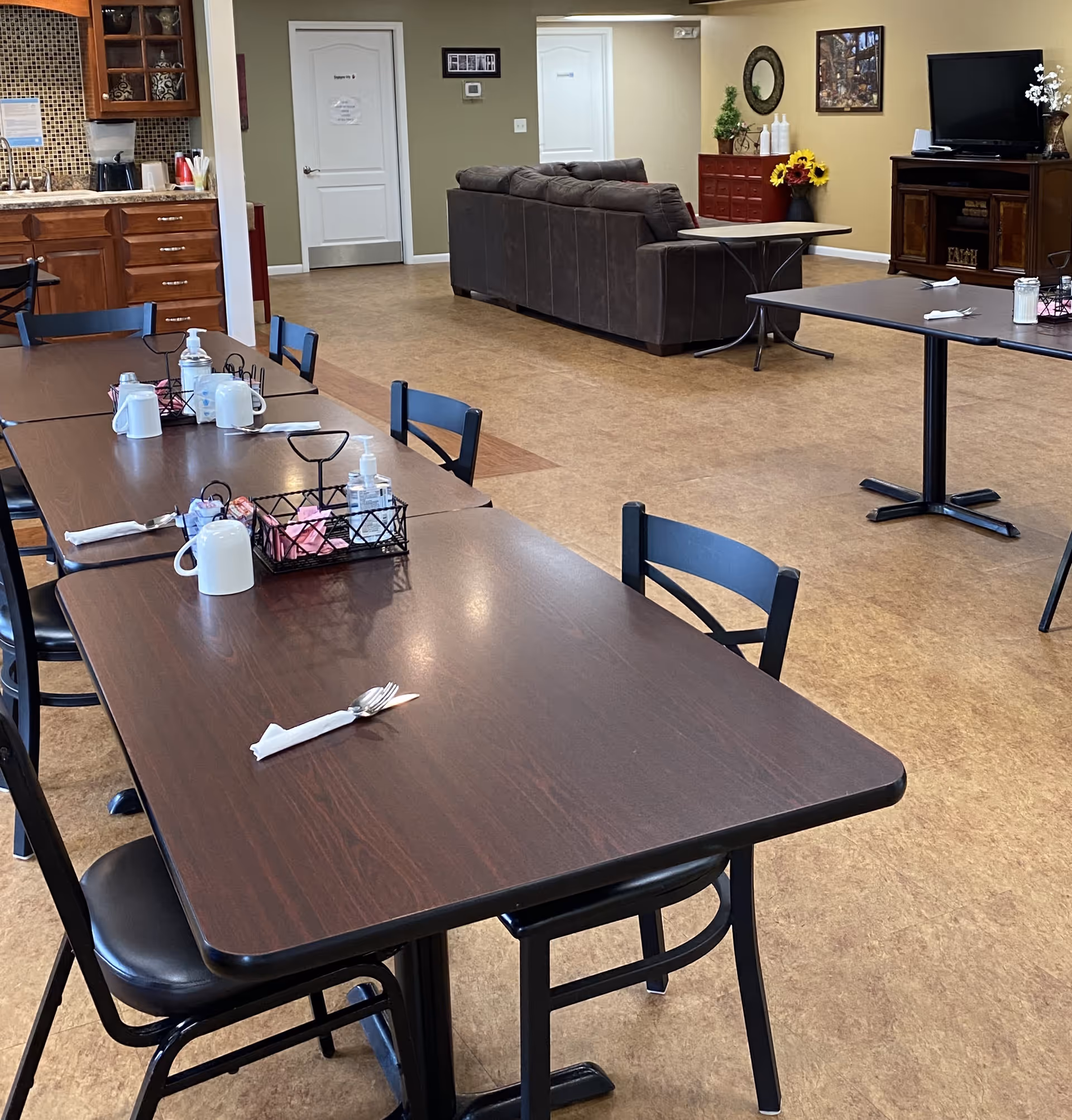 A communal dining and living area in a senior living facility. The foreground shows dark wooden tables with black chairs, set with white mugs, napkins, and utensils. The background features a brown sectional sofa, a TV on a wooden stand, a red cabinet with decorative items, and a kitchen area with wooden cabinets and a coffee maker.
