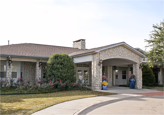 Front exterior view of a single-story stone building with a covered entrance supported by stone pillars. There are bushes and flowers planted along the front, and a curved driveway leads up to the entrance.