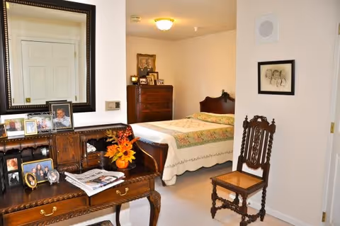 A cozy bedroom with a wooden bed covered in a floral quilt, a wooden dresser with framed photos, a wooden chair with intricate carvings, and a wooden desk with a vase of orange flowers and several framed photographs. A large mirror hangs above the desk, reflecting a closed white door.