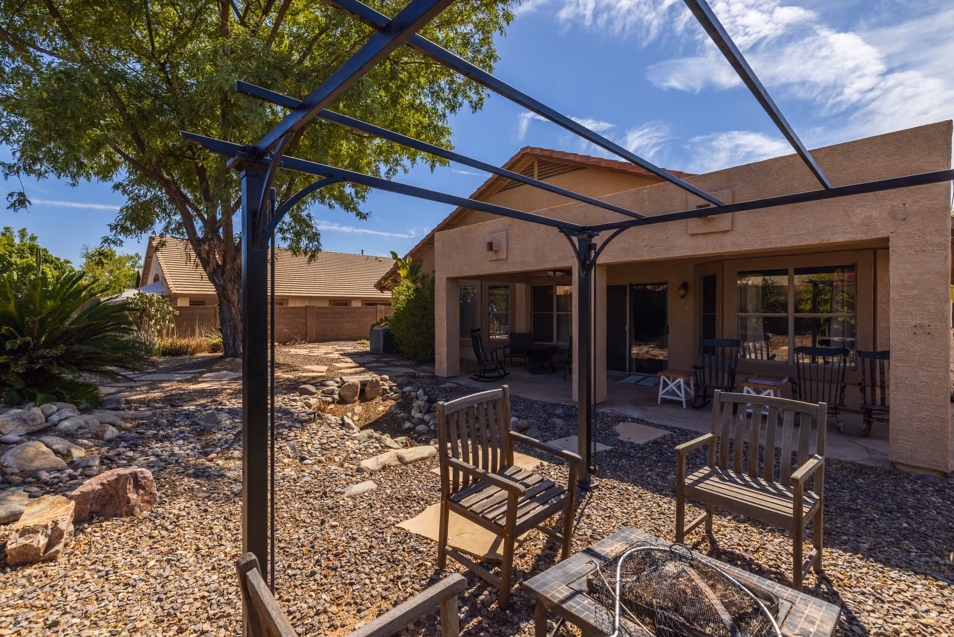 Outdoor patio area with wooden chairs arranged around a fire pit, a metal pergola frame overhead, and a single-story building with a covered porch in the background. The ground is covered with gravel and there are trees and shrubs surrounding the area under a partly cloudy blue sky.