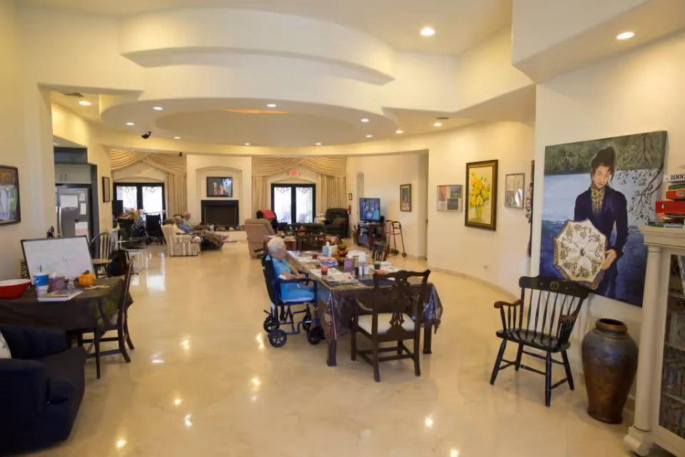 Spacious communal lounge in a senior care home with tables, chairs, seated residents and wall artwork.