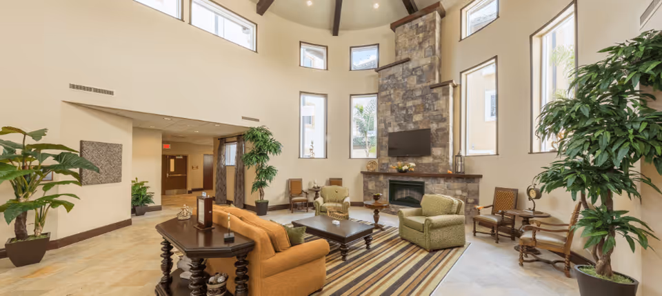 A spacious and well-lit living room area with high ceilings and multiple tall windows. The room features a stone fireplace with a mounted TV above it, a striped area rug, a brown sofa, two green armchairs, several wooden chairs, and potted plants placed around the room.
