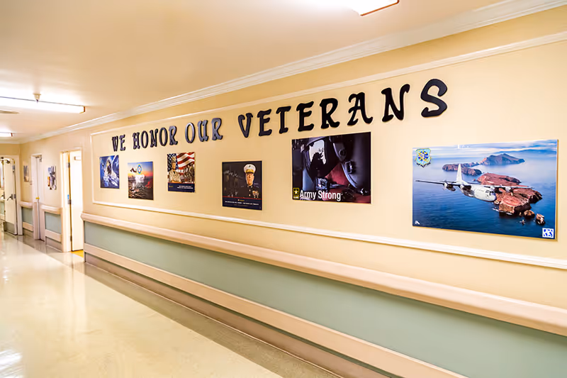 A hallway in a care facility with a display on the wall honoring veterans. The display includes the text 'WE HONOR OUR VETERANS' and several framed pictures related to the military and veterans. The hallway has handrails along the walls and doors leading to rooms.