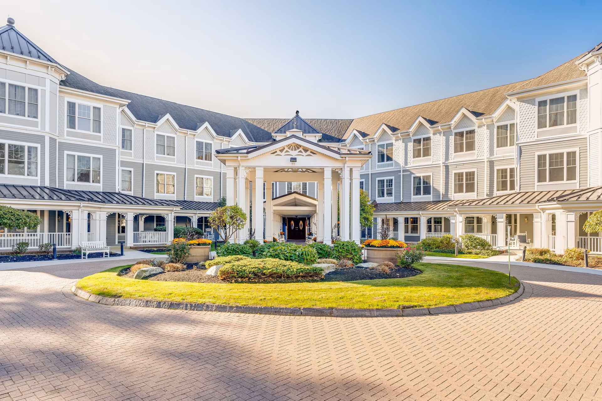 Front exterior view of a large senior living facility building with a circular driveway, landscaped garden with shrubs and flowers, and a covered entrance supported by white columns under a clear sky.