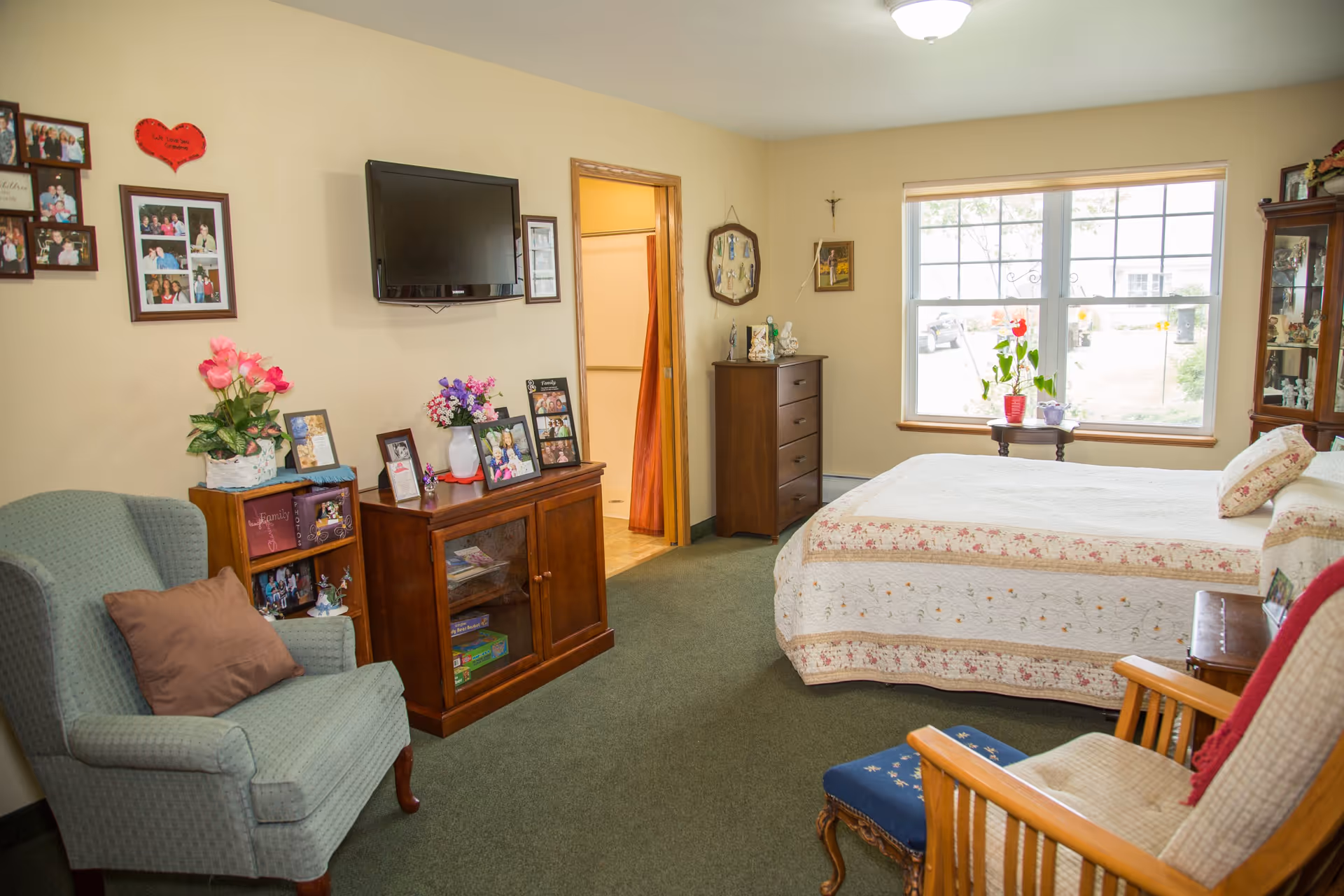 A cozy bedroom in a senior living community with a bed covered in a floral quilt, a large window letting in natural light, a wooden dresser, a TV mounted on the wall, an armchair with a brown pillow, a wooden rocking chair with a footstool, and various framed photos and decorative items on shelves and walls.