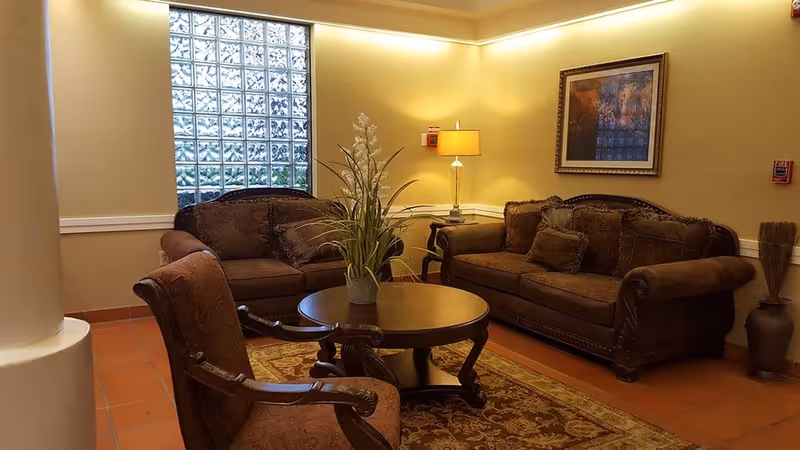 A cozy living room area with two brown upholstered sofas, a matching armchair, a round wooden coffee table with a potted plant on it, a floor lamp, a framed painting on the wall, and a large glass block window letting in natural light.
