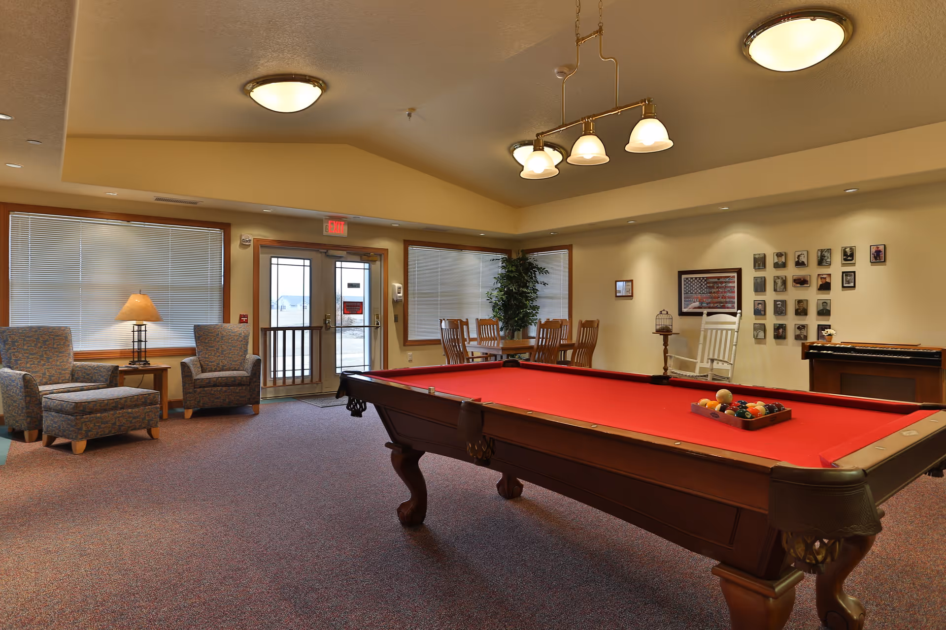 A spacious common room with a red felt pool table in the center, surrounded by comfortable seating including two armchairs with a side table and lamp, a dining table with chairs, a rocking chair, and a piano. The room has large windows with blinds, beige walls decorated with framed pictures, and ceiling lights providing warm illumination.