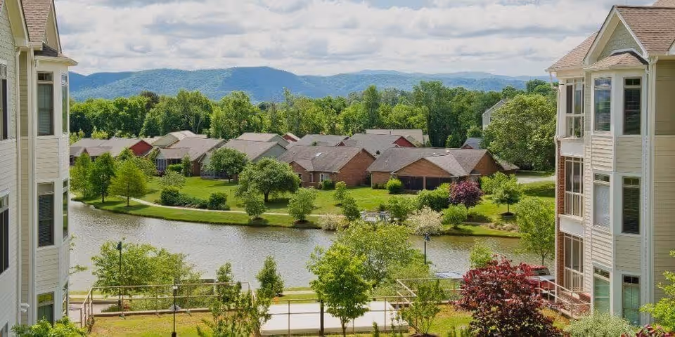 View of a residential community with single-story houses surrounding a small pond, framed by two multi-story buildings on either side, with green trees and mountains in the background under a partly cloudy sky.