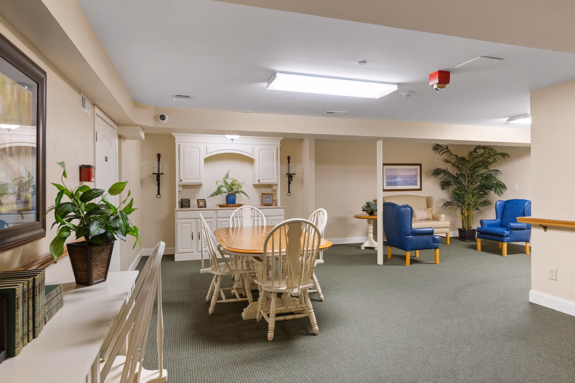 A cozy common area in a senior living facility featuring a wooden dining table with six white chairs, a white cabinet with decorative items and plants, and a seating area with blue and beige armchairs around a small table. The room has beige walls, green carpet, framed artwork, and potted plants.
