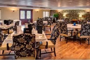 A spacious dining room with multiple tables set for meals, featuring floral patterned chairs, white tablecloths, and neatly arranged table settings. The room has wooden flooring, soft lighting, and a stone accent wall with plants and decorative elements.
