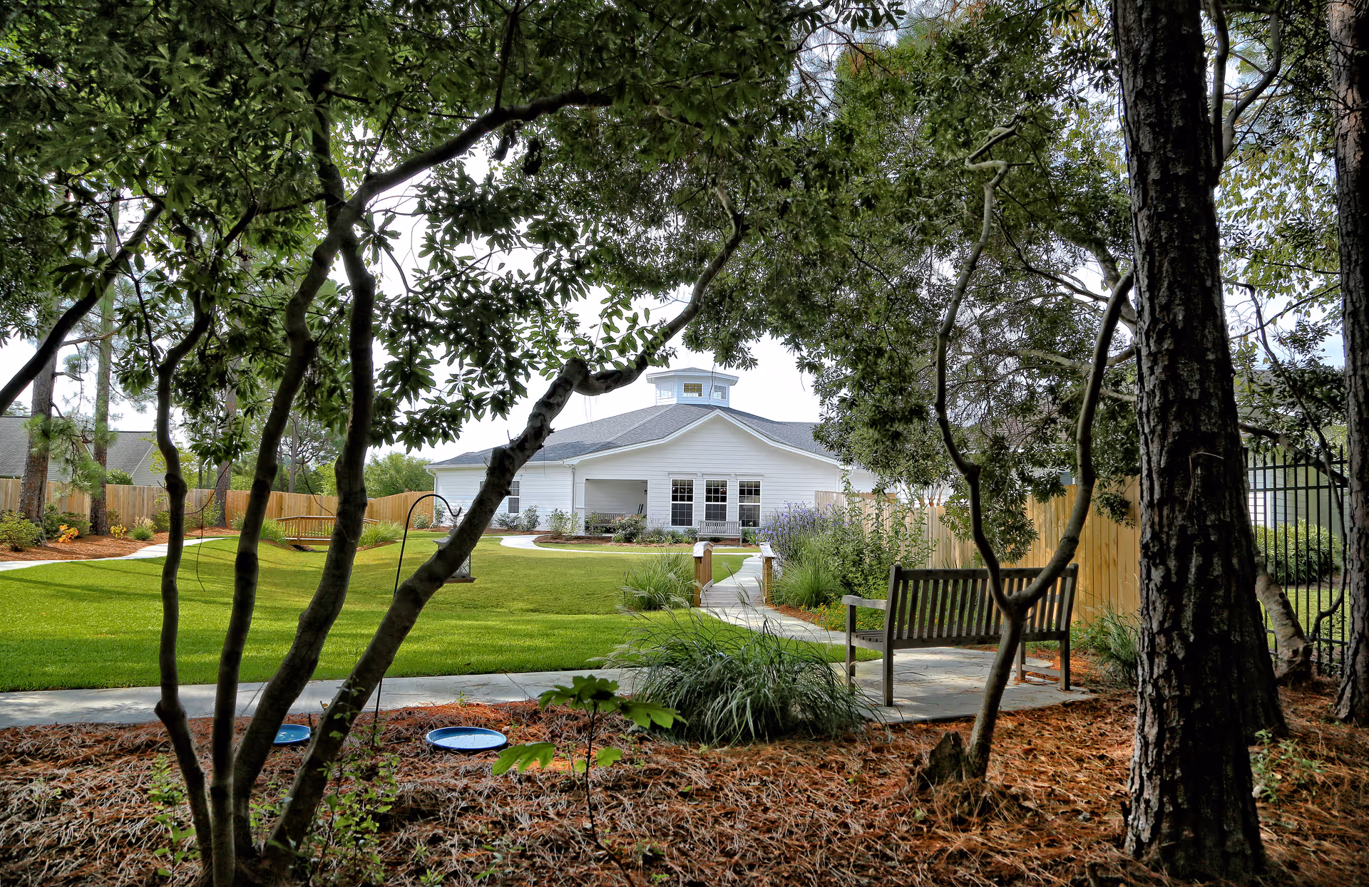 View through trees of a well-maintained garden area with green grass, a wooden bench, and a white building in the background under a cloudy sky.