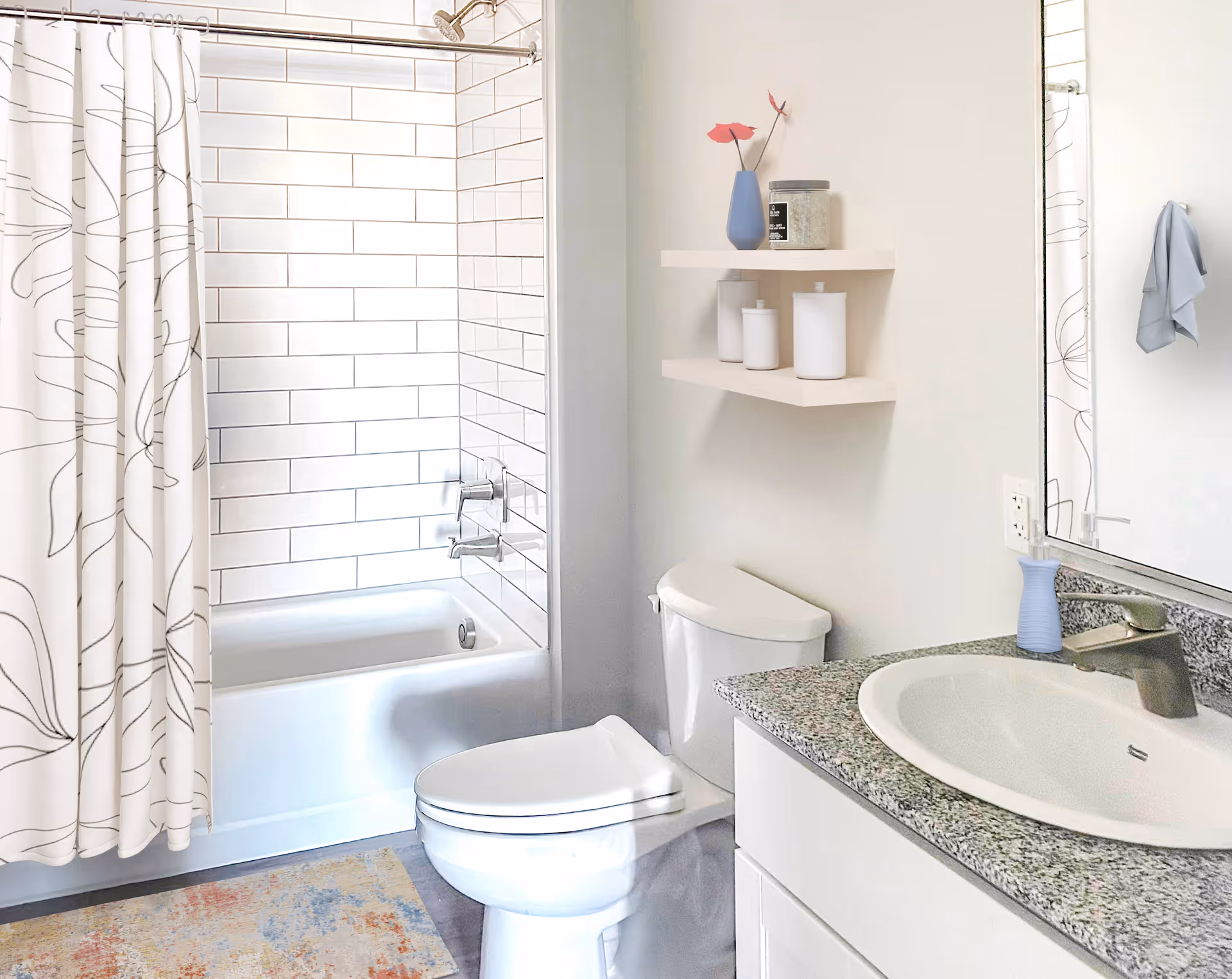 Bright modern bathroom featuring a bathtub/shower with white subway tile, a toilet, sink with granite countertop and mirror, and wall shelves.