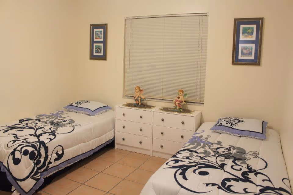 A bedroom with two single beds placed on opposite sides of a white dresser with six drawers. Each bed has a white comforter with black and gray floral patterns and a matching pillow. On top of the dresser are two small angel figurines. The room has beige walls with two framed pictures hanging on either side of a window with closed blinds.