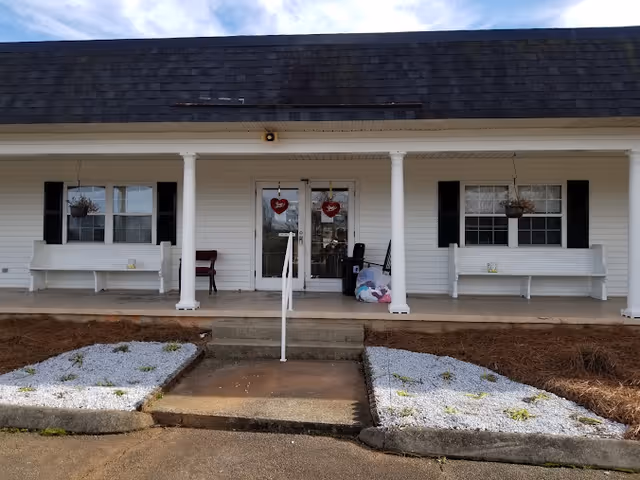 Front porch of a building with white siding and black shutters, featuring two white benches on either side of a double glass door entrance decorated with red heart-shaped ornaments. There are hanging flower baskets above each bench and a small set of concrete steps leading up to the porch with a white handrail in the center. The ground in front of the porch has two rectangular garden beds filled with white gravel and some sparse greenery.