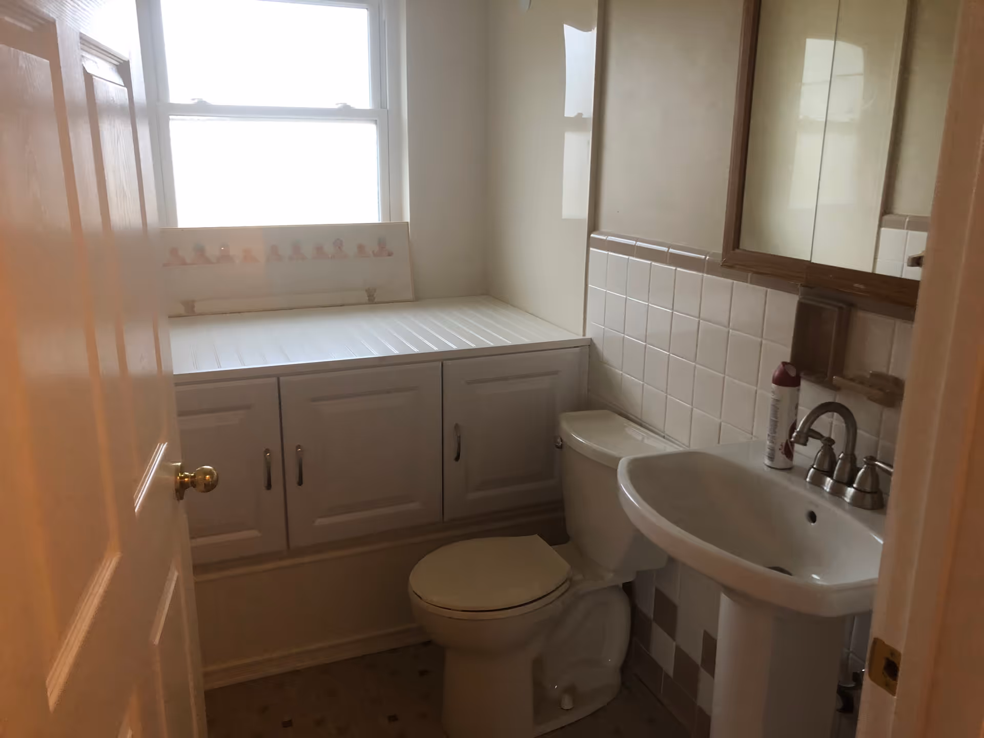 A small bathroom with a white toilet and pedestal sink. There is a tiled backsplash behind the sink and toilet, and a wooden framed mirror above the sink. A window with frosted glass is above a white cabinet with multiple doors. A can of air freshener is placed on the sink.
