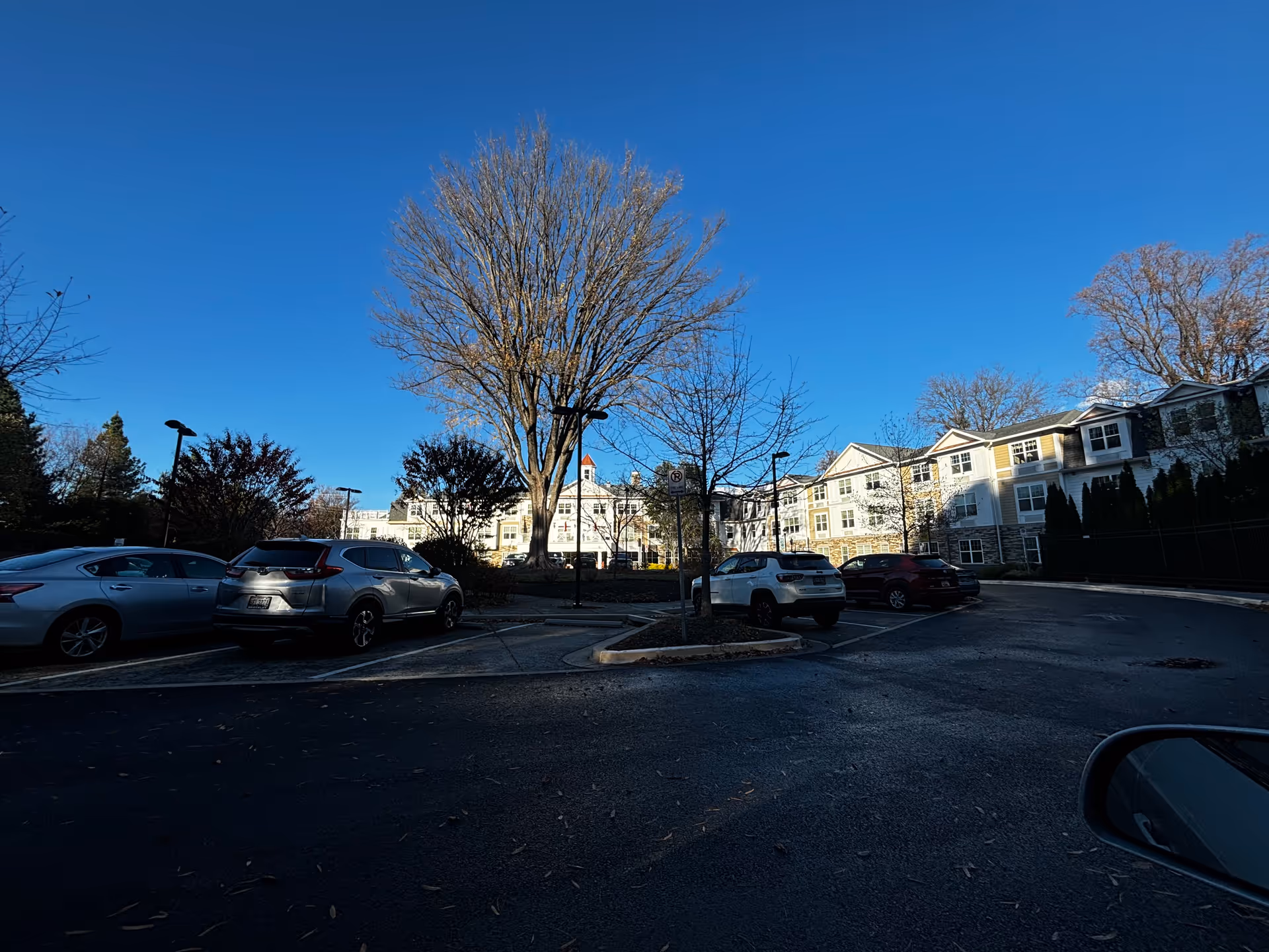 Parking lot with several parked cars in front of a multi-story senior living facility building under a clear blue sky. Leafless trees and some bushes are visible around the parking area.