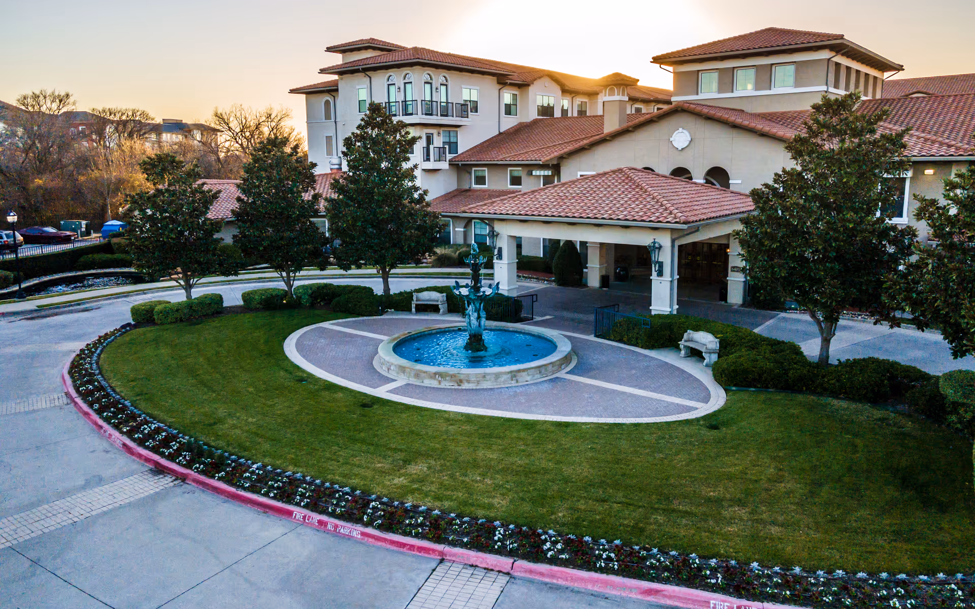 Exterior view of Conservatory At Plano senior living facility at sunset, showing a circular driveway with a central fountain, manicured lawn, trees, and a multi-story building with red-tiled roofs and balconies.