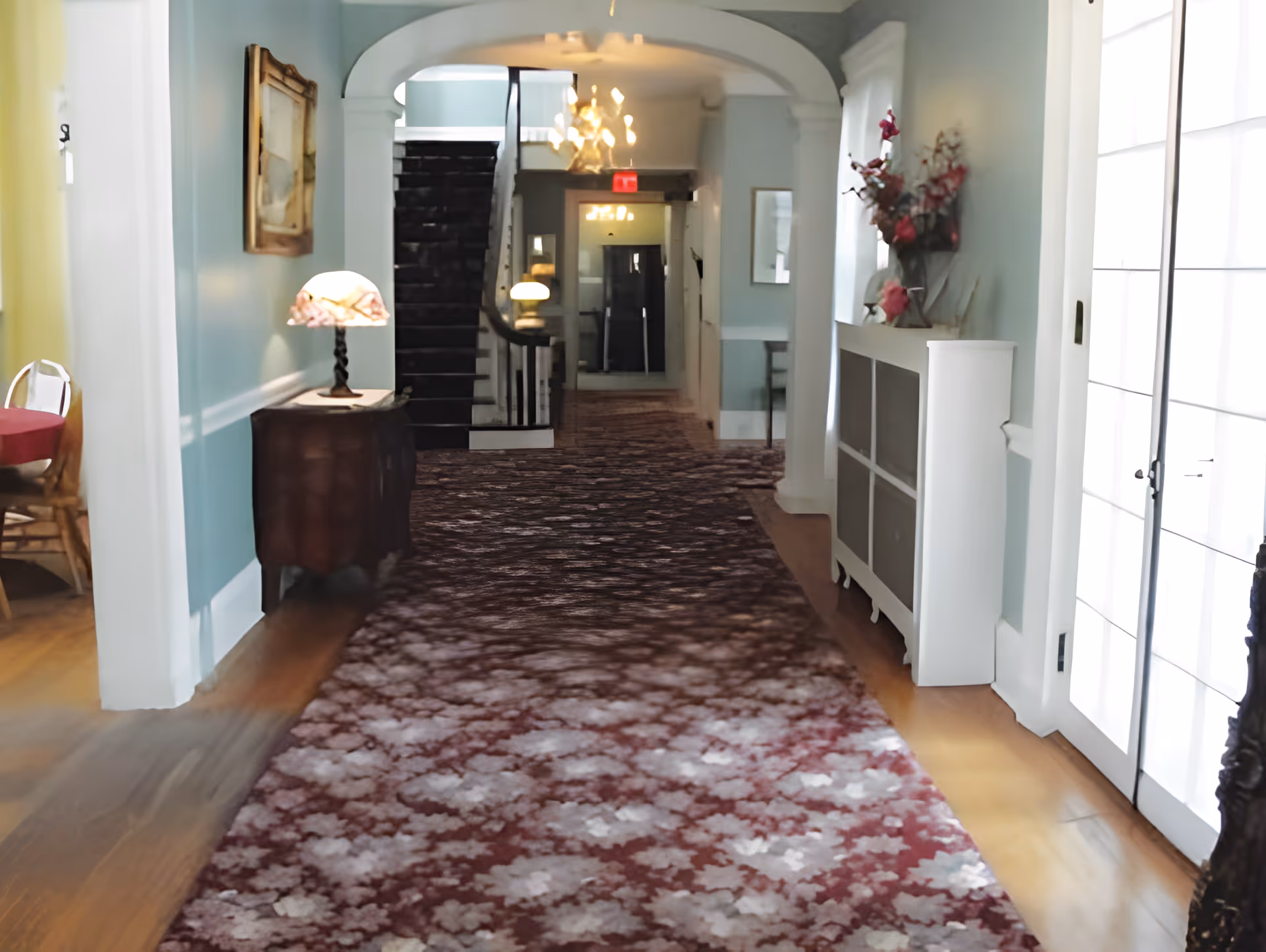 A carpeted interior hallway with a patterned runner, side tables and lamps, a white cabinet with flowers, and a staircase at the far end.