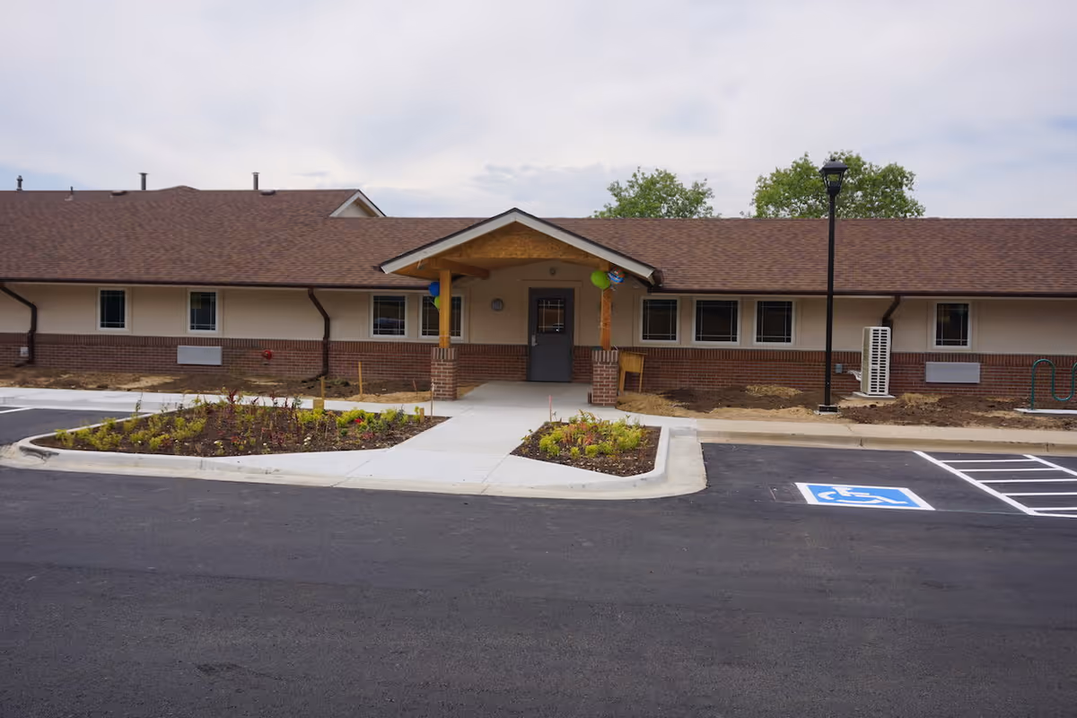 Exterior view of a single-story assisted living facility building with a brown roof and beige walls with brick accents. There is a covered entrance with wooden posts, a sidewalk leading to the door, landscaped flower beds on either side, a parking lot with a handicapped parking space, and a street lamp near the entrance.