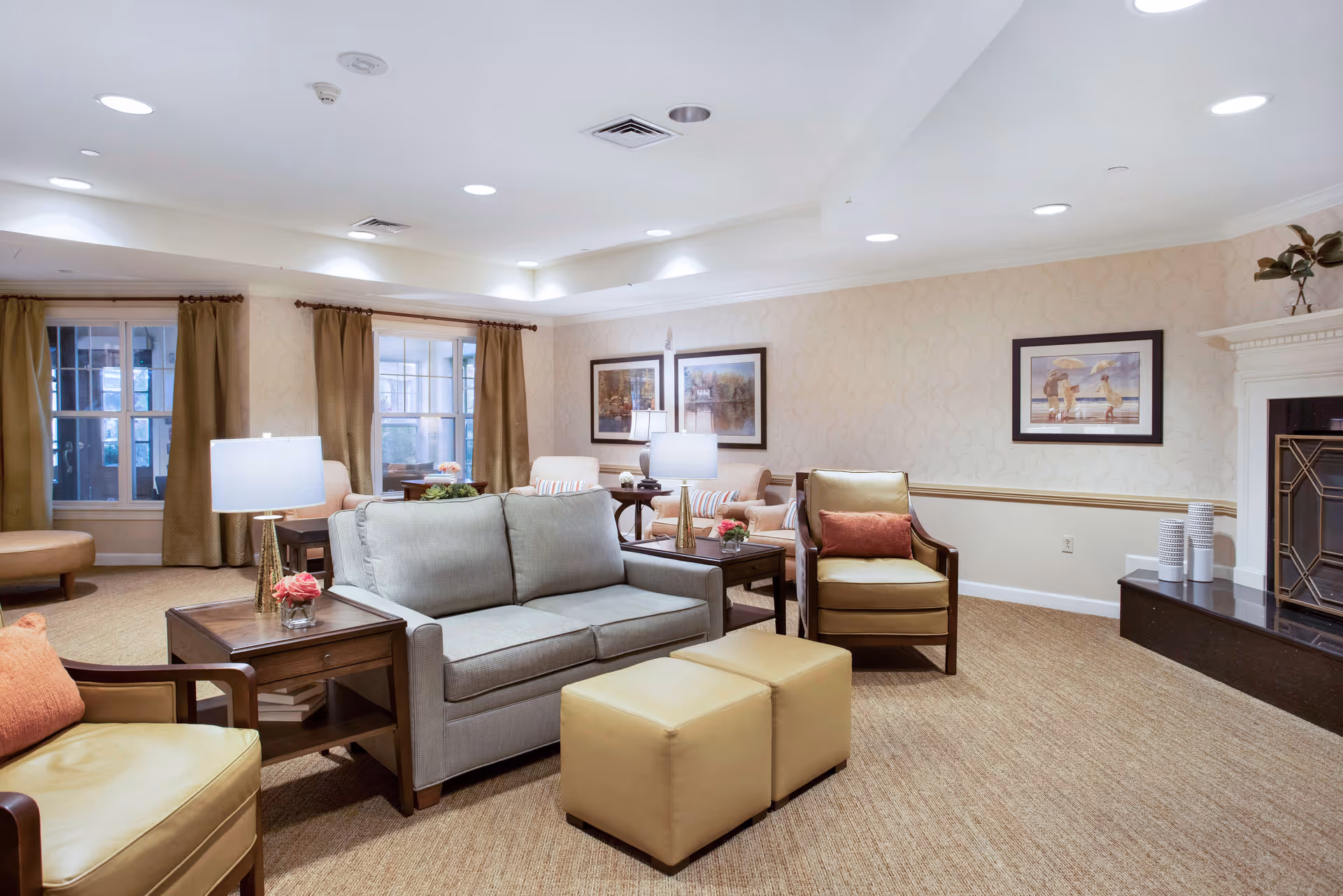 A cozy living room with beige walls and carpet, featuring a gray sofa, two tan armchairs with orange cushions, two beige ottomans, wooden side tables with lamps, framed artwork on the walls, and a fireplace with decorative vases on the mantel.