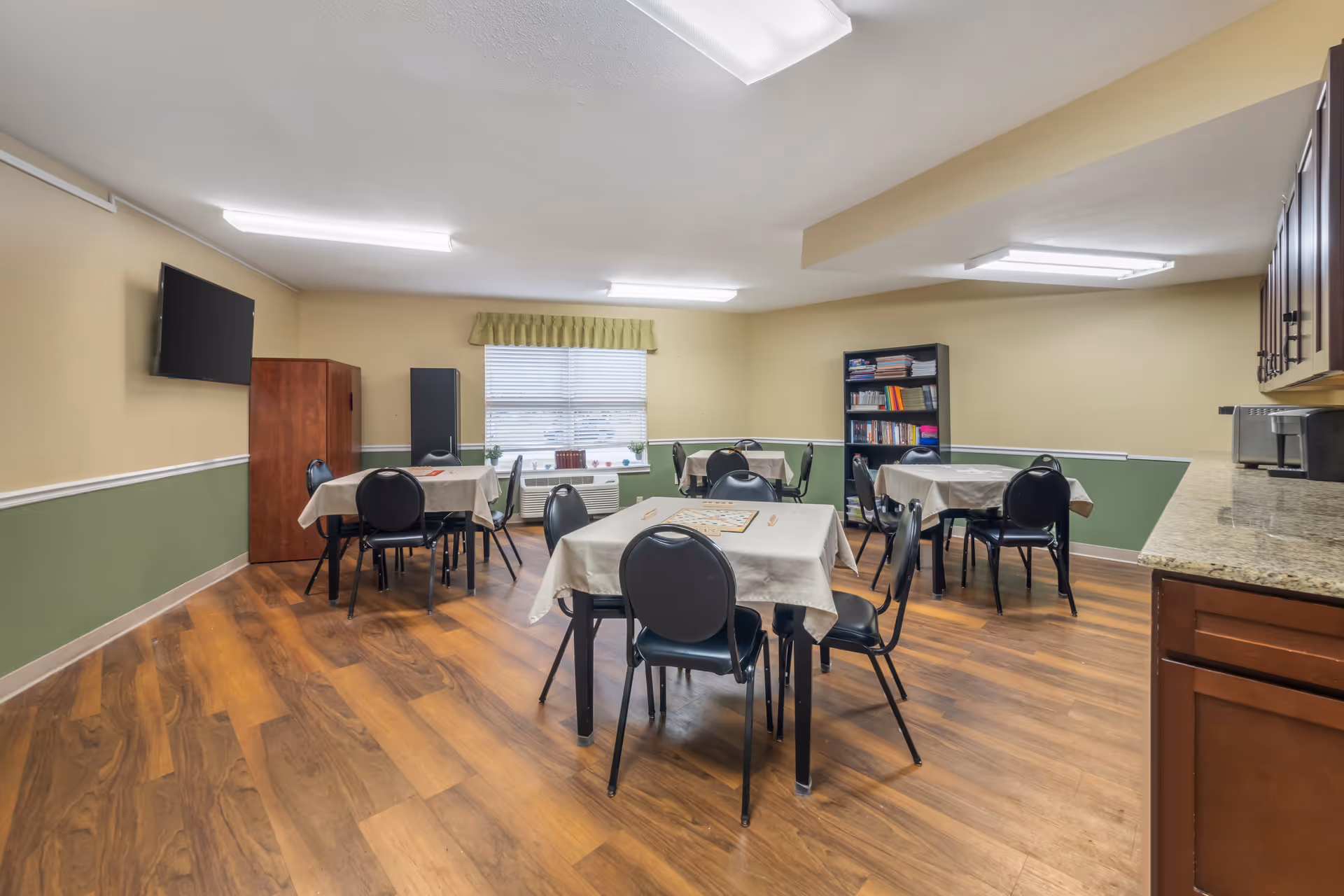 A well-lit room with several tables covered with beige tablecloths, each surrounded by black chairs. The room has wood flooring, beige and green walls, a window with blinds and a green valance, a mounted flat-screen TV, a wooden cabinet, a black bookshelf filled with books and games, and a countertop with kitchen appliances on the right side.