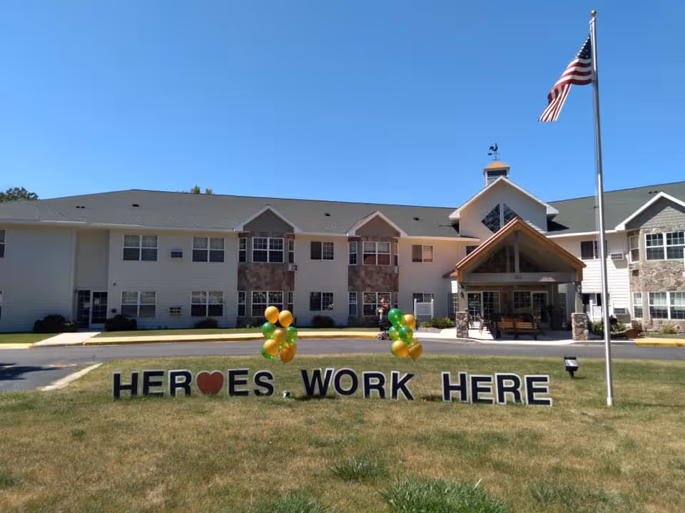 Front exterior view of a senior living facility building with a green lawn in front. On the lawn, large letters spell out 'HEROES WORK HERE' with a red heart symbol replacing the letter O in HEROES. There are green and gold balloons attached to the letters. An American flag flies on a flagpole to the right of the sign. The building has multiple windows, a peaked roof, and a covered entrance.