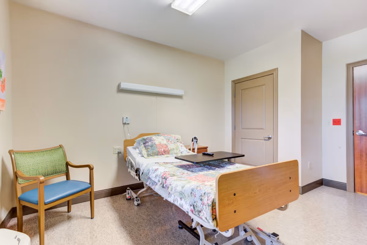 A small, simple room with a hospital-style bed covered with a floral quilt, a green and blue chair, a wooden door, and a small overbed table with a remote control on it. The walls are light beige and the floor is a light-colored linoleum.