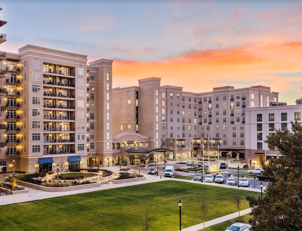 Exterior view of The Barclay at SouthPark senior living facility at sunset, showing a large multi-story building with balconies, lit windows, a covered entrance, parked cars, and a well-maintained lawn in the foreground.