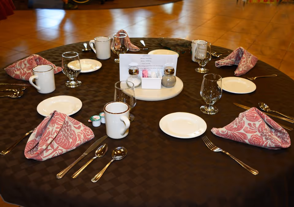 A round dining table set for six with a dark checkered tablecloth, white plates, cups, water glasses, silverware, and pink patterned cloth napkins folded neatly. In the center, there is a rotating tray with salt and pepper shakers, sugar packets, and a small sign.