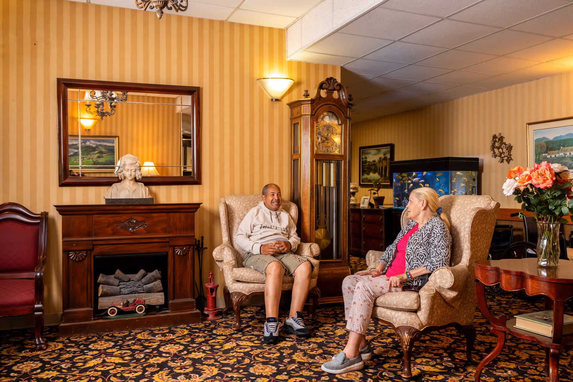 Two elderly people sitting and conversing in a warmly decorated living room with patterned carpet, striped wallpaper, a wooden fireplace with a bust sculpture, a large wall mirror, a grandfather clock, an aquarium, and a table with a vase of flowers.