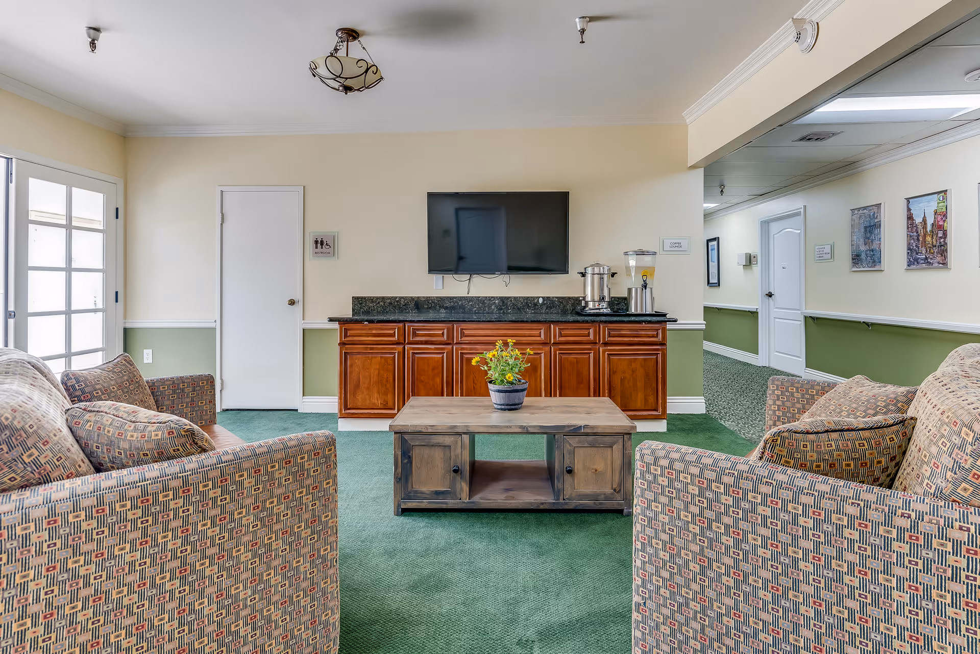 A senior living facility common area with two patterned armchairs facing a wooden coffee table with a potted plant. Behind the table is a wooden cabinet with a black countertop, holding a beverage dispenser and a coffee urn. A flat-screen TV is mounted on the wall above the cabinet. To the left, there is a door labeled restroom, and to the right, a hallway with framed pictures on the walls.
