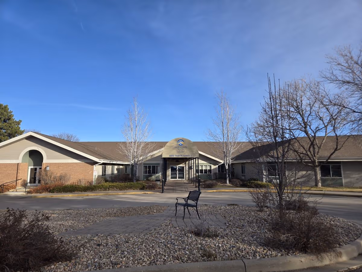 Front exterior of a single-story senior living building with an entrance canopy, circular driveway, bench, and leafless trees under a blue sky.