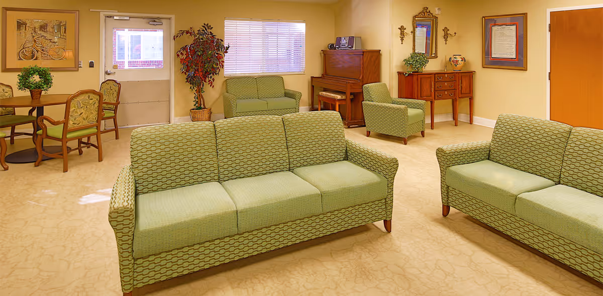 A cozy living room area in a senior living facility with two green patterned sofas, a matching armchair, a wooden piano, a small wooden table with a plant, a round table with four chairs, framed artwork on the walls, and a door with a window letting in natural light.