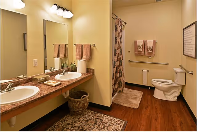 A senior living community bathroom with two sinks on a granite countertop, a large mirror above the sinks, a wicker basket underneath, a toilet with grab bars, a shower with a floral curtain, and towels hanging on the wall. The floor is wooden with a patterned rug near the sinks and a bath mat near the shower.