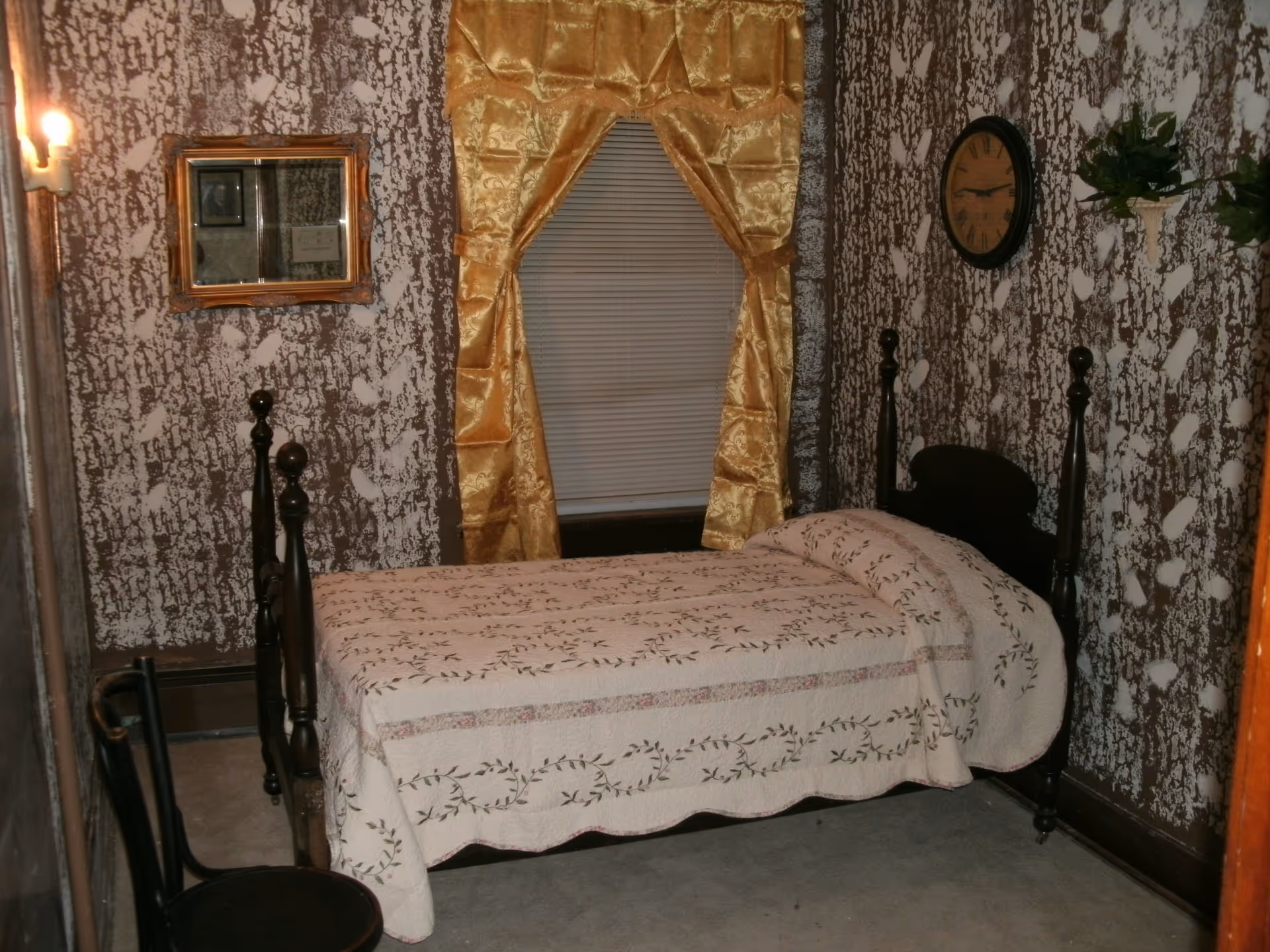 A small bedroom with a single dark wooden bed covered with a white quilt featuring a floral pattern. The walls have a textured brown and white pattern. A window with closed blinds is adorned with gold satin curtains. A round wall clock and two small green plants in white wall-mounted planters are on the right wall. A wooden chair is positioned near the foot of the bed, and a small framed mirror hangs on the left wall.